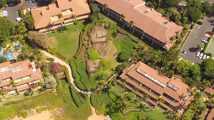 Aerial view of the fishing heiau site at Makena Surf: a rocky, overgrown knoll between two buildings, with the path to the beach curving around its base